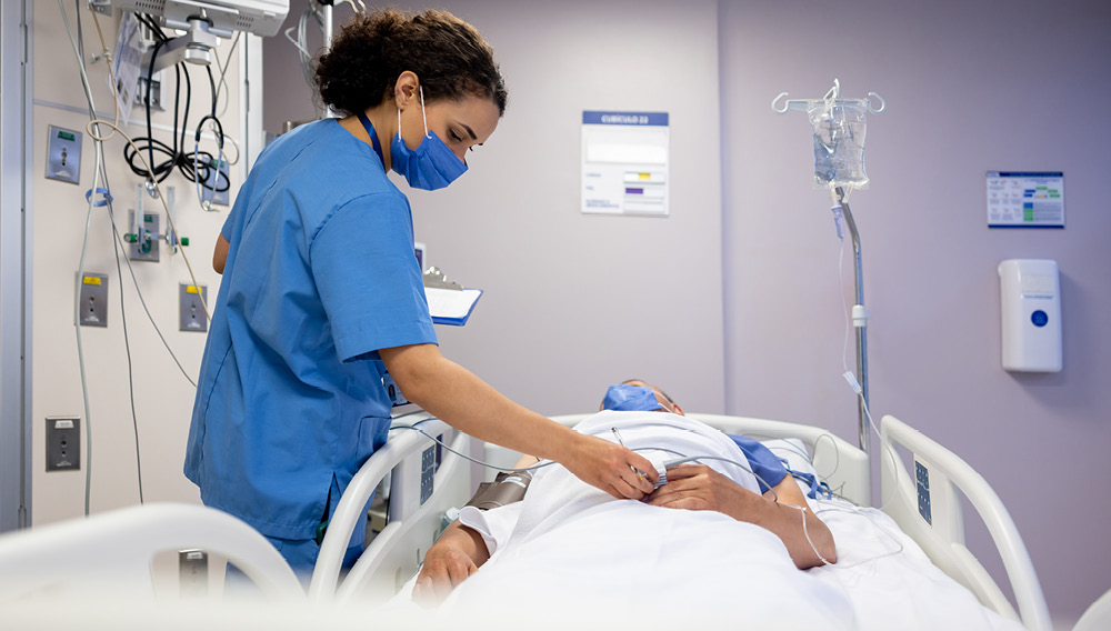 Masked nurse in ICU room checking the pulse oximeter on a patient’s finger