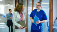 Hospital administrator carrying a laptop speaking with a nurse holding a folder