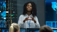 Woman in button-down shirt and lanyard addressing a group of analysts seated before computers