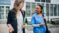 Pair of women in public health and nursing, respectively, walking in a hospital courtyard side by side