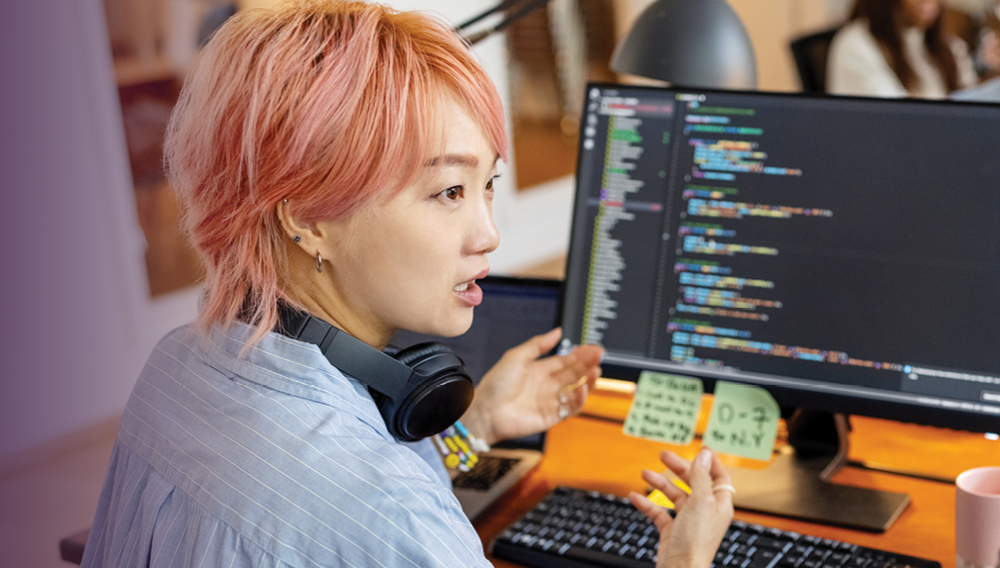 Person sitting at a computer, with headphones around their neck, talking to someone