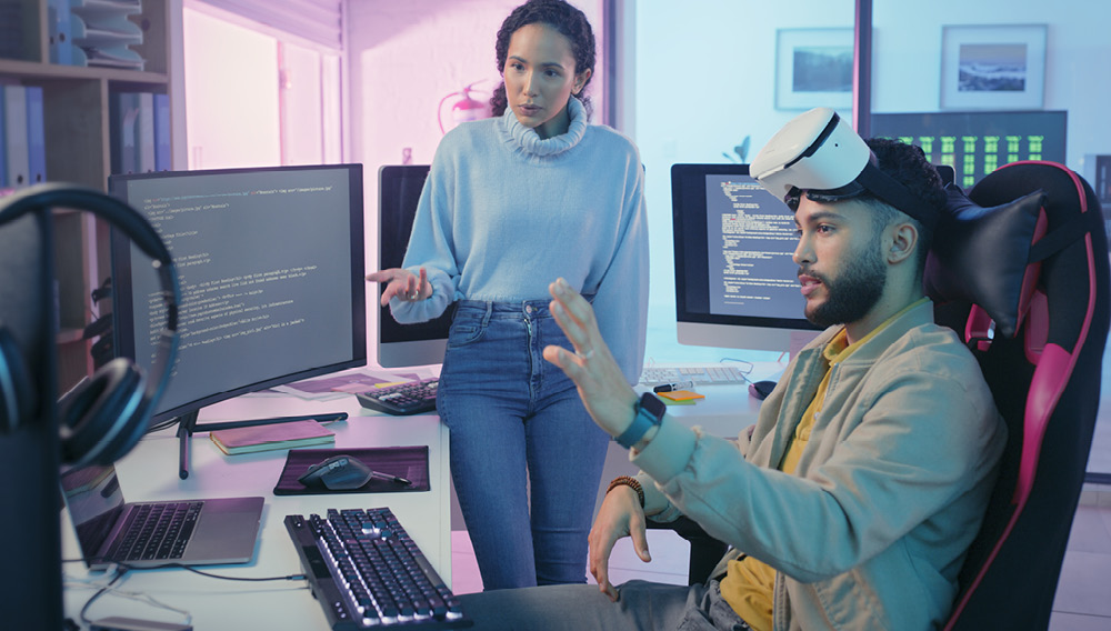 Man seated at computer desk with VR goggles pulled up on his forehead, explaining game design to a co-worker