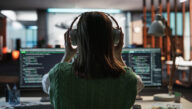 View from behind of person positioning headphones on their ears as they study two screens of code